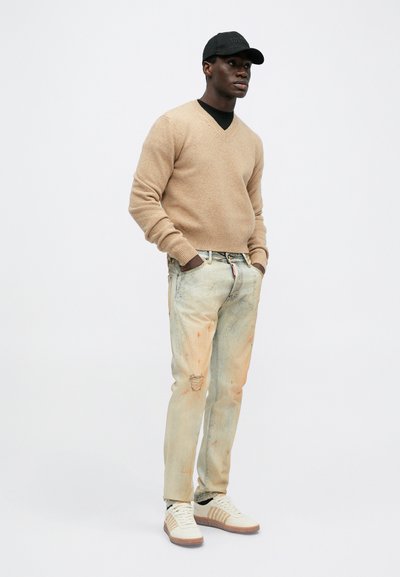 Beiger Strickpullover mit V-Ausschnitt, hellblaue Jeans mit leichter Ausbleichung und Farbspritzern sowie weiße Sneakers mit braunen Akzenten und Streifen.