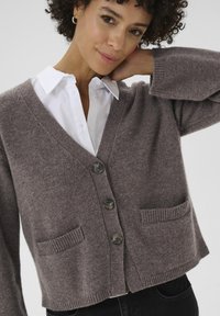 Brązowy sweter z wełny z dekoltem w serek, trzema guzikami i przednimi kieszeniami. Żebrowane mankiety i dół oraz miękka tekstura. Połączony z białą koszulą.