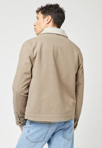 Giacca marrone, con texture e un colletto morbido di colore chiaro. Il design include cuciture visibili e un dettaglio con bottone sul polsino.