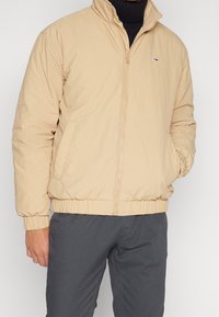 Beige bomber bunda na zip s vysokým límcem, pružnými manžetami a lemem, se malým logem na levé hrudi. Nositá přes tmavě šedé kalhoty.