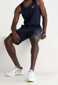 Débardeur bleu marine et short avec logo Nike ; chaussures de sport blanches avec accents néon ; tissu lisse et léger ; pose assise.