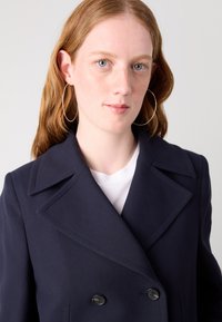 Manteau double boutonnage bleu marine avec grands revers, doté de boutons noirs. Associé à un t-shirt blanc, des cheveux longs et ondulés, et des boucles d'oreilles en argent.