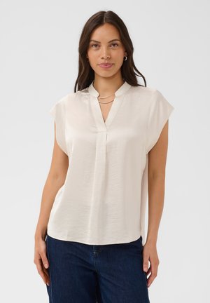 Jeune femme aux cheveux foncés portant un chemisier beige à manches courtes et un jean bleu foncé, debout devant un fond clair uni.