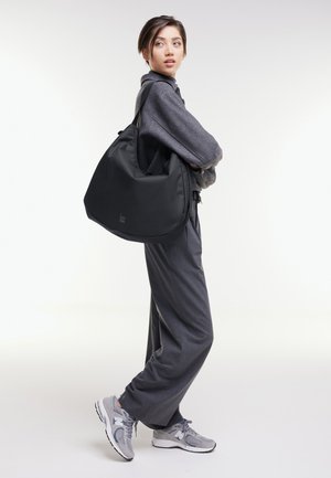 Junge Frau in grauem Outfit und Turnschuhen trägt eine große schwarze Schultertasche und schaut über ihre Schulter vor einem einfarbigen Hintergrund.