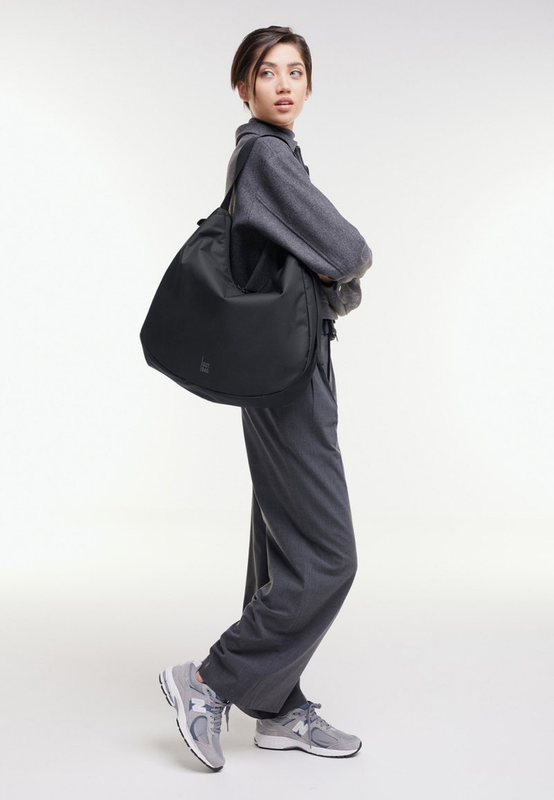 Junge Frau in grauem Outfit und Turnschuhen trägt eine große schwarze Schultertasche und schaut über ihre Schulter vor einem einfarbigen Hintergrund.