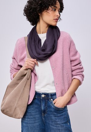 Žena v světle růžovém strukturovaném saku, bílé košili, tmavě fialovém šátku, modrých džínách a držící velkou béžovou kabelku přes rameno.