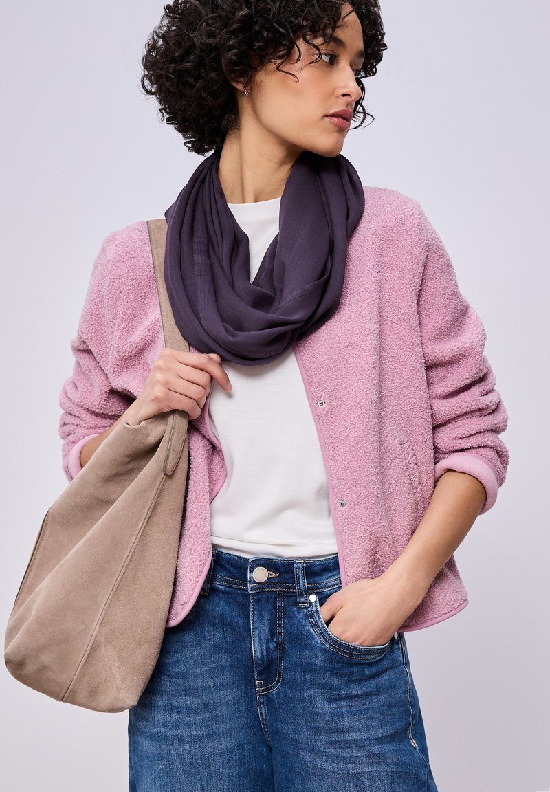 Femme portant une veste texturée rose clair, une chemise blanche, une écharpe violet foncé, un jean bleu et tenant un grand sac à bandoulière beige.