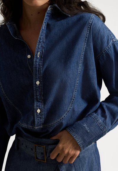 Sieviete, kas valkā tumši zilu džinsa kreklu ar pogām un saskaņotu augsta vidukļa jostu, roku pielāgojot jostas sprādzi.