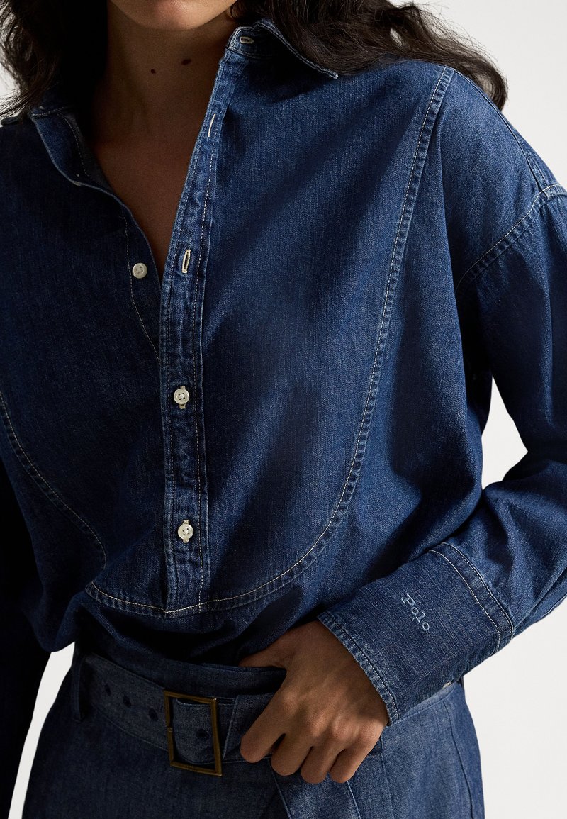 Donna che indossa una camicia di jeans blu scuro con bottoni e una cintura coordinata a vita alta, mentre si regola la fibbia della cintura con la mano.