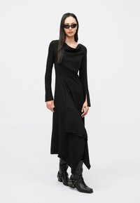 Μαύρο μακρύ φόρεμα με λαιμόκοψη καζάκα, μακριά μανίκια, ασύμμετρο τελείωμα και ραμμένες λεπτομέρειες. Συνδυασμένο με μαύρες μπότες μέχρι το γόνατο και γυαλιά ηλίου.