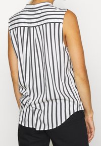 Blusa senza maniche con colletto, caratterizzata da righe verticali bianche e nere, tessuto morbido e orlo arrotondato sul retro.