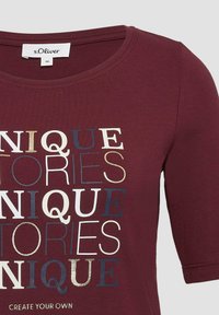 Bordeaux T-Shirt mit kurzen Ärmeln und rundem Ausschnitt, mit mehrfarbigen Textgrafiken in Gold, Weiß und Blau. Weiches Baumwollmaterial.