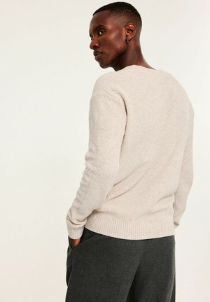 Vaaleanbeige neulepusero, jossa on teksturoitu malli, resorit hihansuissa ja helmassa, yhdistetty tummanharmaisiin housuihin, takakatsaus.