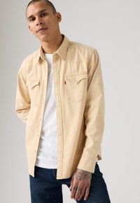 Mann i beige skjorte med knapper over hvit t-skjorte og mørke jeans, som viser en tatovert hånd med hodeskalle- og stjernemotiver.