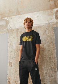 Sort bomulds T-shirt med et gult grafiktryk og PUMA-logoet, sammen med sorte træningsbukser, der viser PUMA-emblemet.