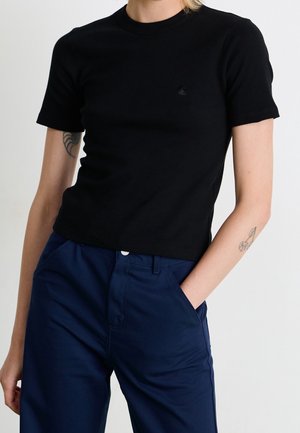 Person wearing a fitted black short-sleeve top and high-waisted dark blue pants with one hand in pocket, showing small arm tattoos.