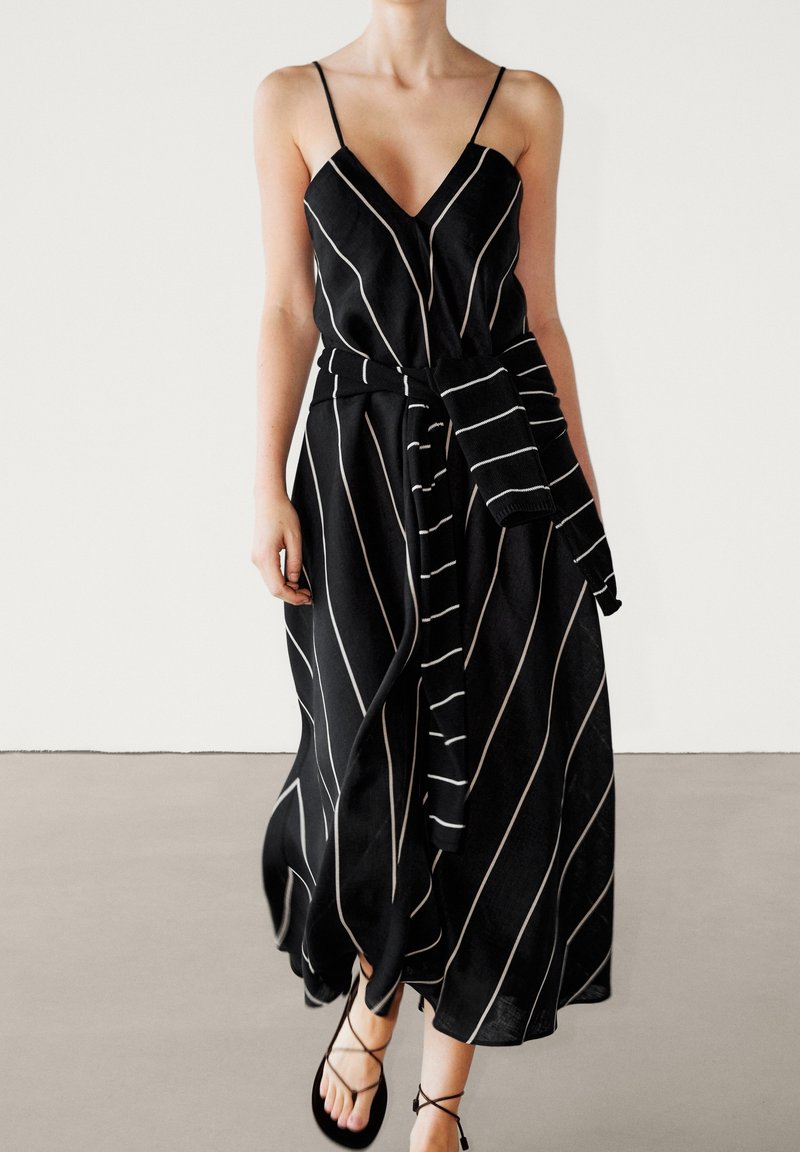 Robe maxi noire avec fines rayures blanches verticales. Présente un col en V, des bretelles spaghetti et une ceinture à nouer, fabriquée dans un tissu léger.