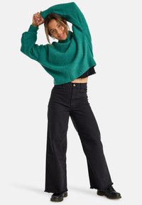 Turkusowy, oversize'owy sweter z dzianiny o wysokim dekolcie, zestawiony z czarnymi spodniami o wysokim stanie i szerokich nogawkach z surowymi brzegami oraz czarnymi skórzanymi butami.