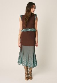 Femme portant une robe marron sans manches en maille avec un bord volanté bleu et un ourlet plissé, associée à des bottes montantes à motifs et des bracelets dorés.