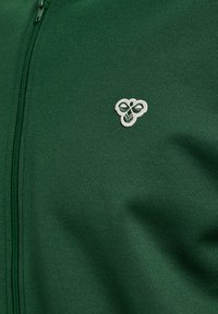 Tummanvihreä kangas edessä vetoketju ja pieni valkoinen kirjailtu mehiläislogo vetoketjun lähellä oikealla puolella.