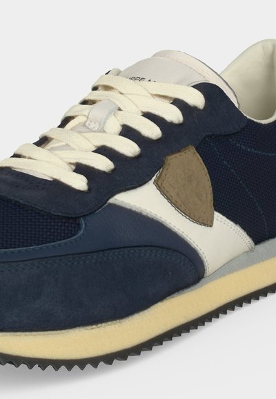 Gros plan d'une basket bleu marine et blanche avec des lacets crème, des textures en suède et en mesh, un patch latéral beige et une semelle beige à rainures.
