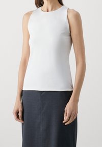 Vit ärmlös topp i slät tyg, med rund halsringning och en figursydd siluett, uppvisad med en mörk blyertskjol.