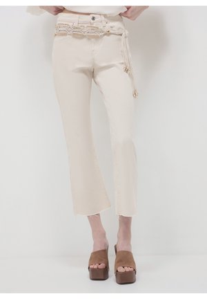 Femme portant un pantalon crème court droit avec des ourlets effilochés, une ceinture tressée perlée et des sandales marron à plateforme à bout ouvert.