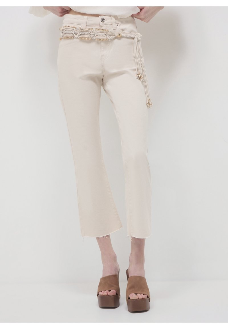 Femme portant un pantalon crème court droit avec des ourlets effilochés, une ceinture tressée perlée et des sandales marron à plateforme à bout ouvert.