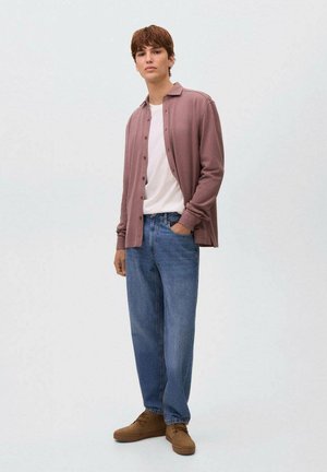 Mlad moški stoji z eno roko v žepu, oblečen v mauve srajco s knofci nad belim majico, modre kavbojke in rjave čevlje na enobarvni podlagi.