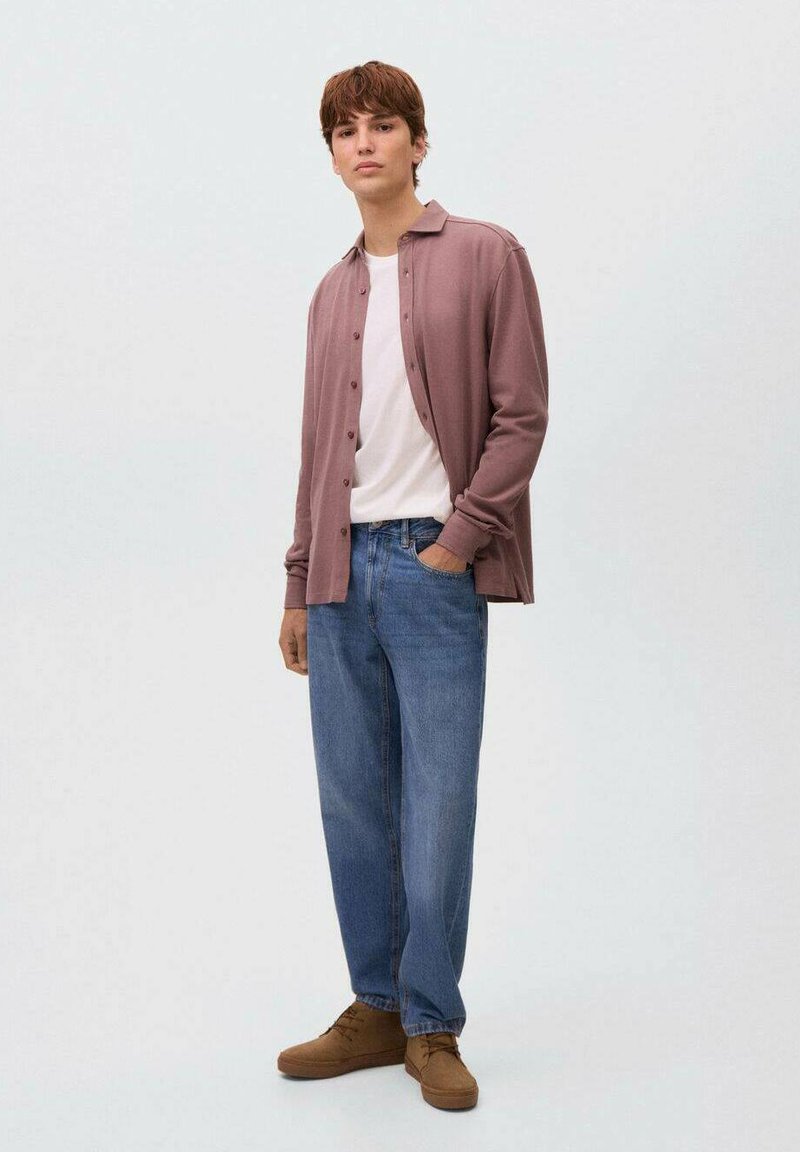 Giovane uomo in piedi con una mano in tasca, che indossa una camicia mauve con bottoni sopra una maglietta bianca, jeans blu e scarpe marroni su uno sfondo neutro.
