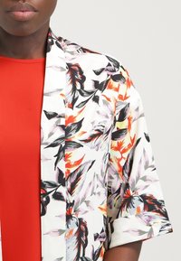 Květinová bunda ve stylu kimona s bílým základem a živými červenými, černými a fialovými květinovými vzory. Měkká, lehká látka s krátkými rukávy.