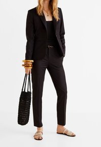 Zwarte fitted blazer, bijpassende broek en een zwart geweven tas. Accessoires zijn onder andere gouden armbanden en beige sandalen met een platte zool.