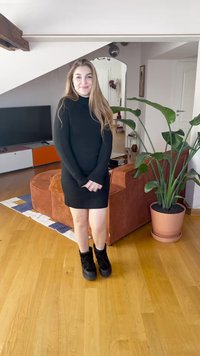 Juoda struktūruota golfų tipo suknelė ilgomis rankovėmis, derinama su juodais platformos batais. Vidaus interjeras su dideliu žaliu augalu ir baldais.