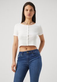 Vit kort knäppt skjorta med korta ärmar, ribbad struktur, med en liten broderad logotyp. Bärs med blå jeans med hög midja.