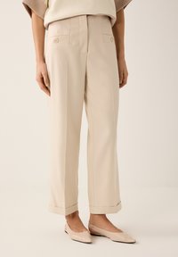 Beige op maat gemaakte broek met twee voorzakken, lichte plooien bij de taille en opgerolde pijpen, gecombineerd met platte beige schoenen.