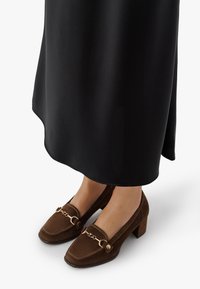 Fötter i bruna mocka loafers med klack och guldkedjedetalj, i kombination med en lång svart kjol på vit bakgrund.