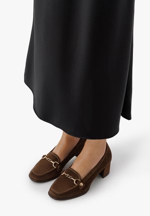 Feet wearing brown suede block-heeled loafers with gold chain detail, paired with a long black skirt on white background.
