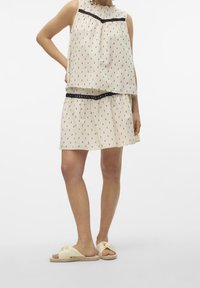 Haut sans manches crème superposé sur une jupe assortie, avec de petits motifs noirs et une bordure noire à la taille. Porté avec des sandales de couleur claire.