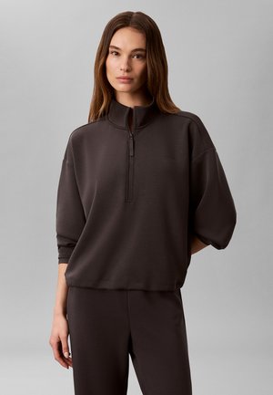 Kvinde med glat brunt hår iført en mørkebrun half-zip pullover og matchende bukser mod en ensartet lys grå baggrund.