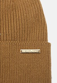 Brun strikket hue med ribstrikket tekstur og en guldmetallogo på forsiden.