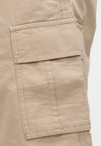 Τσέπη μπεζ υφασμάτινου παντελονιού cargo με καπάκι και εμφανή ραφή στο πλευρό του ενδύματος.