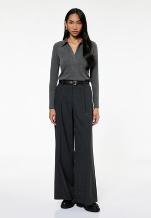 Mujer con el cabello negro y largo, vistiendo una camisa gris oscuro de manga larga con cuello, pantalones negros anchos con cinturón y zapatos negros, de pie.