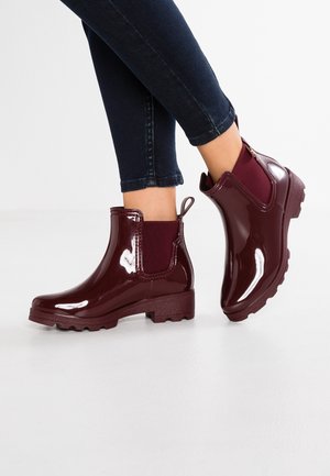 Glinsterende donkerrode enkellaarzen voor regen met elastische zijkanten, gedragen met donkergetinte jeans op een witte ondergrond.