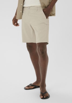Mand iført beige shorts, en lys skjorte og sorte klipklappere, stående foran en ensfarvet baggrund.