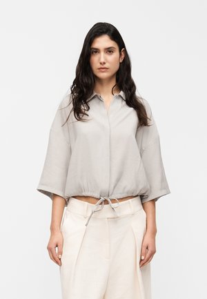 Graues, kurz geschnittenes Hemd mit Knopfleiste am Kragen, weiten Ärmeln und Kordelzug am Saum, kombiniert mit hellfarbigen, plissierten Hosen.