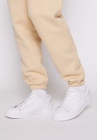 Baskets blanches avec une tige en cuir lisse, bout arrondi et semelle en caoutchouc texturée. Portées avec un pantalon de jogging beige resserré aux chevilles.