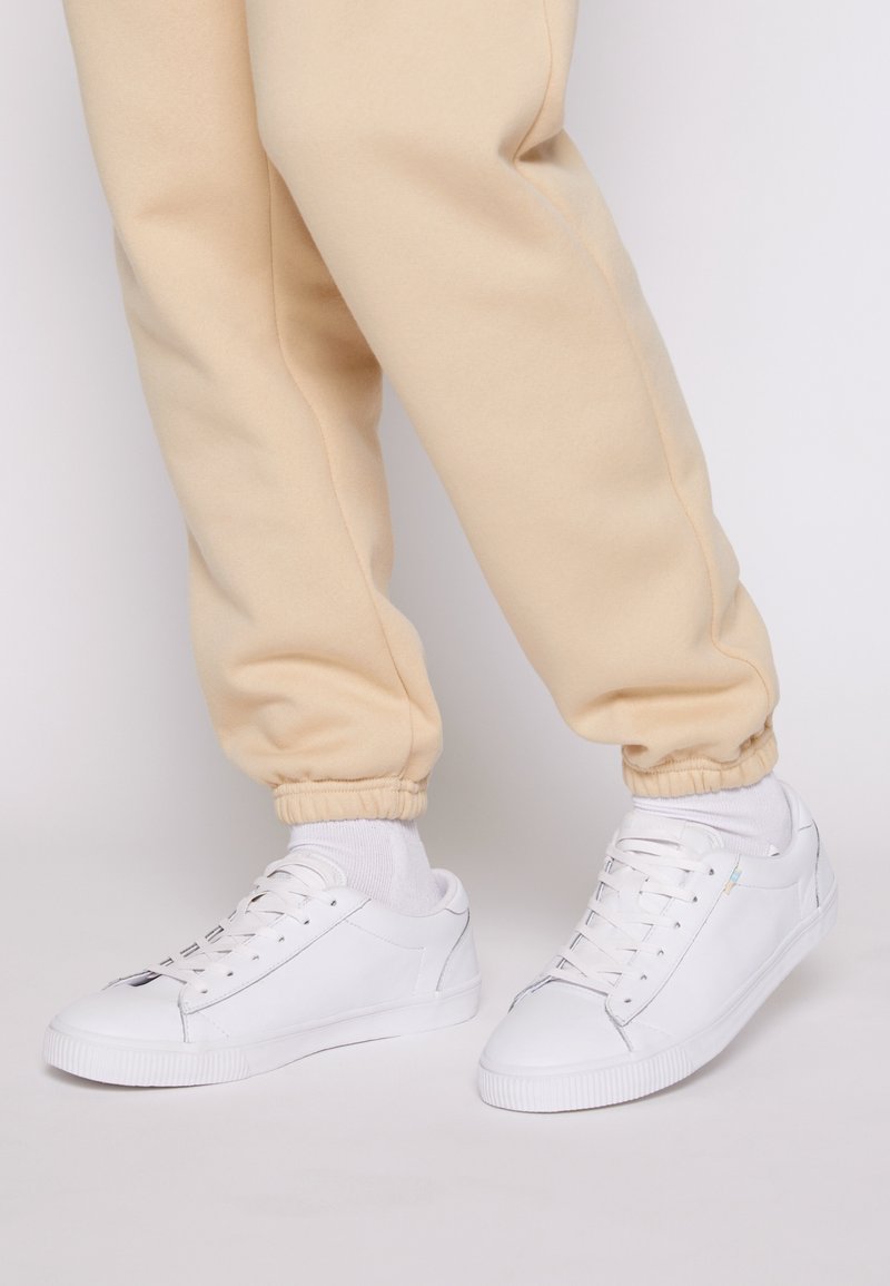 Baskets blanches avec une tige en cuir lisse, bout arrondi et semelle en caoutchouc texturée. Portées avec un pantalon de jogging beige resserré aux chevilles.