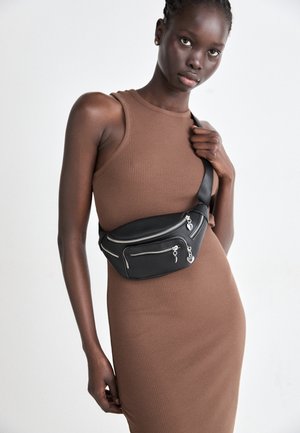 Femme portant une robe marron sans manches à côtes et un sac ceinture en cuir noir avec plusieurs poches zippées sur la poitrine.