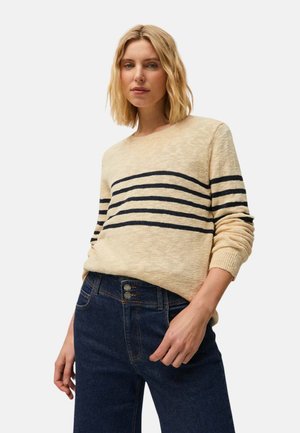 Mujer rubia con suéter beige con rayas negras horizontales y vaqueros azul oscuro de talle alto, de pie con una mano en el bolsillo.