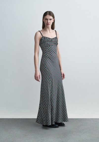 Γυναίκα που φοράει μακρύ, αμάνικο μαύρο φόρεμα με λευκά και κόκκινα γεωμετρικά σχέδια και μαύρα παπούτσια, στέκεται μπροστά σε απλό φωτεινό γκρι φόντο.